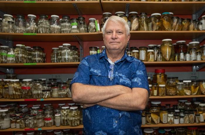 A man in a blue shirt poses in front of jars of biological samples.